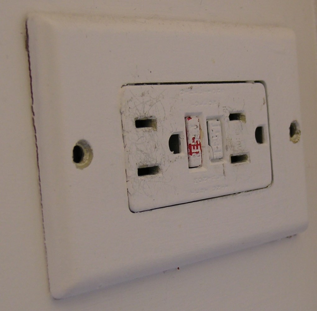 A light-colored wall with a ground-fault 
circuit interrupter installed. It shows that it was painted; one can see some 
of the original red peeking through the paint on the TEST button. 